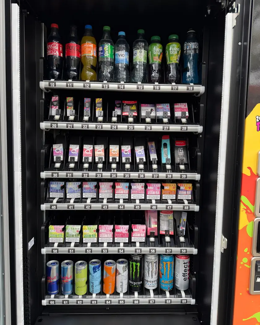 Geöffneter TRALLAFITTI-Automat: oben gekühlte Getränke, darunter Vapes und Zubehör in nummerierten Fächern, unten Energy-Drinks